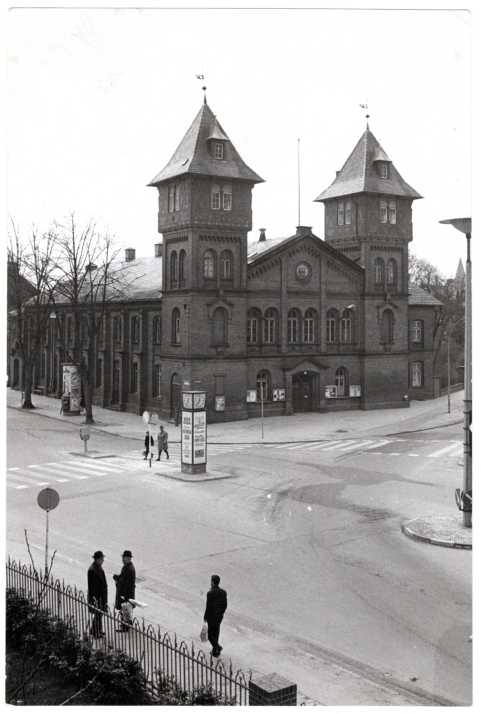 MTV-Halle, Lindenstraße 30 in Lüneburg, etwa 1965. Privatbesitz Beckmann | ArGW. Es ist ein schwarz-weißes Bild. Aus einer erhöhten Position aufgenommen zeigt es eine Straßenkreuzung. Auf der gegenüberliegenden Seite steht die Turnhalle mit zwei markanten eckigen Türmen. Sie ist prachtvoll mit Backstein gemauert.